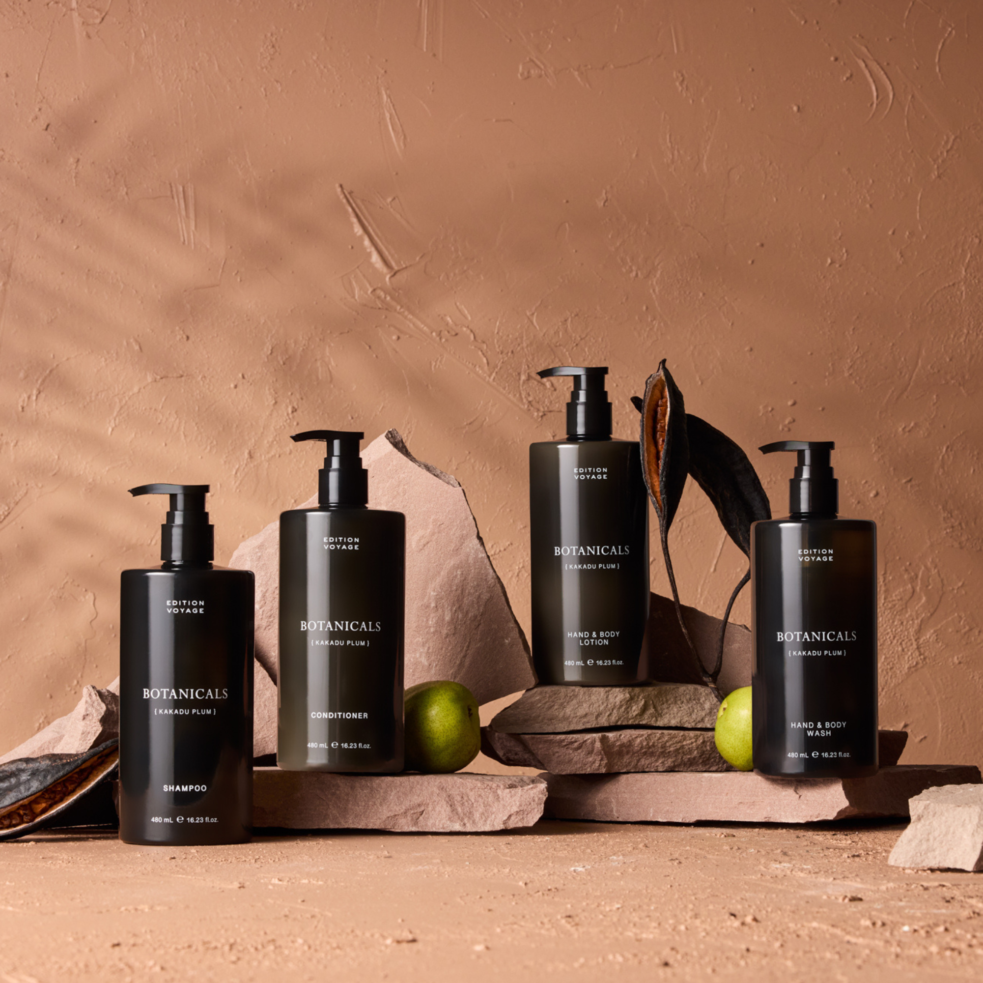 Four black Edition Voyage Botanicals toiletry bottles displayed on stone slabs with green fruits.